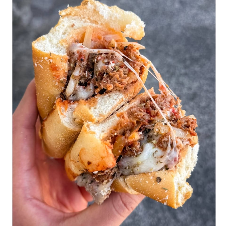 Hand holding a toasted sandwich filled with shredded meat, melted cheese, onions, and crispy toppings.