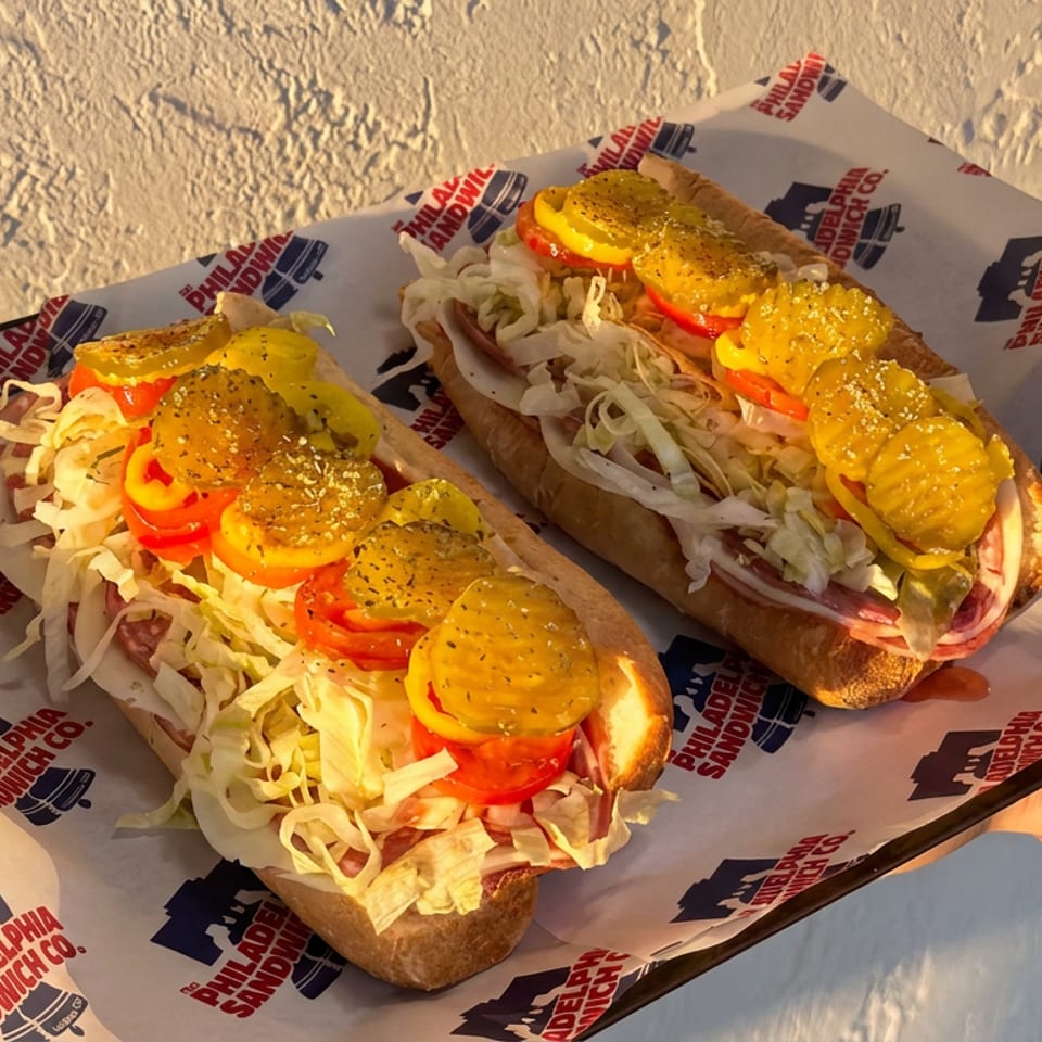 Two loaded sandwiches topped with shredded lettuce, tomatoes, pickles, and seasoning, served on branded paper in warm sunlight.