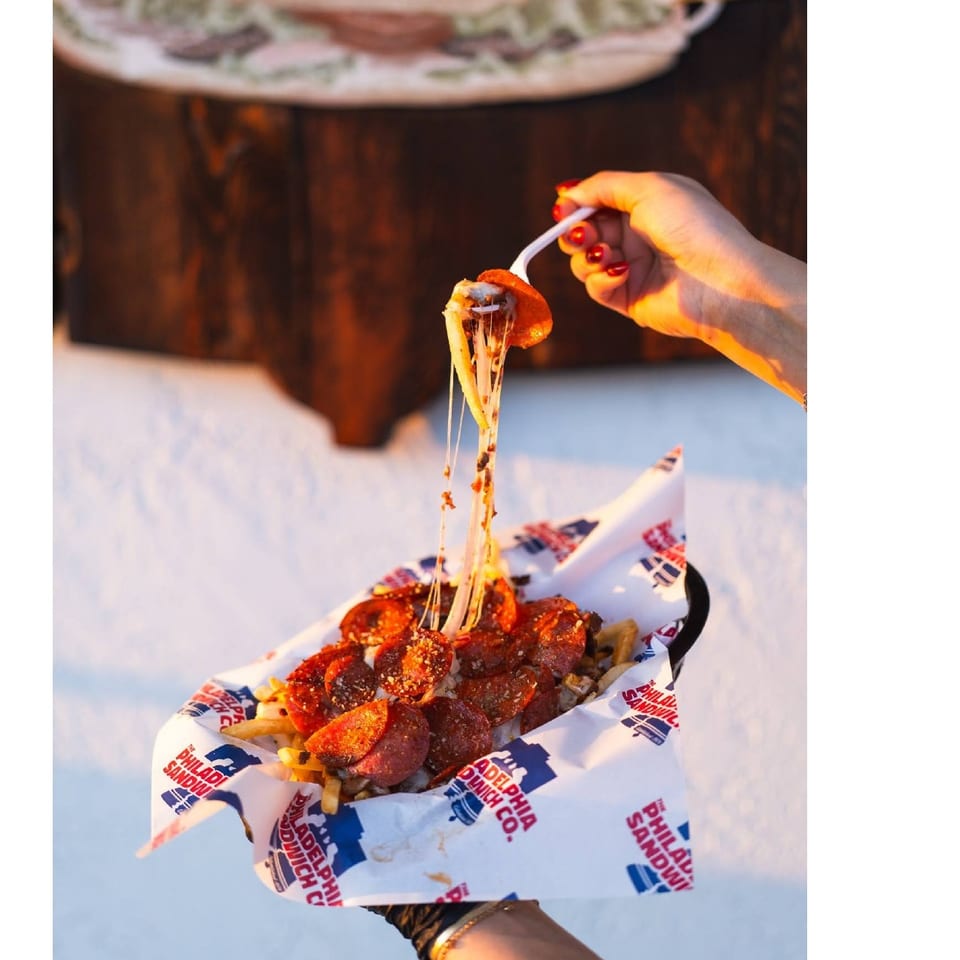 Hand lifts a forkful of melted cheese from pepperoni-covered fries served in branded paper.