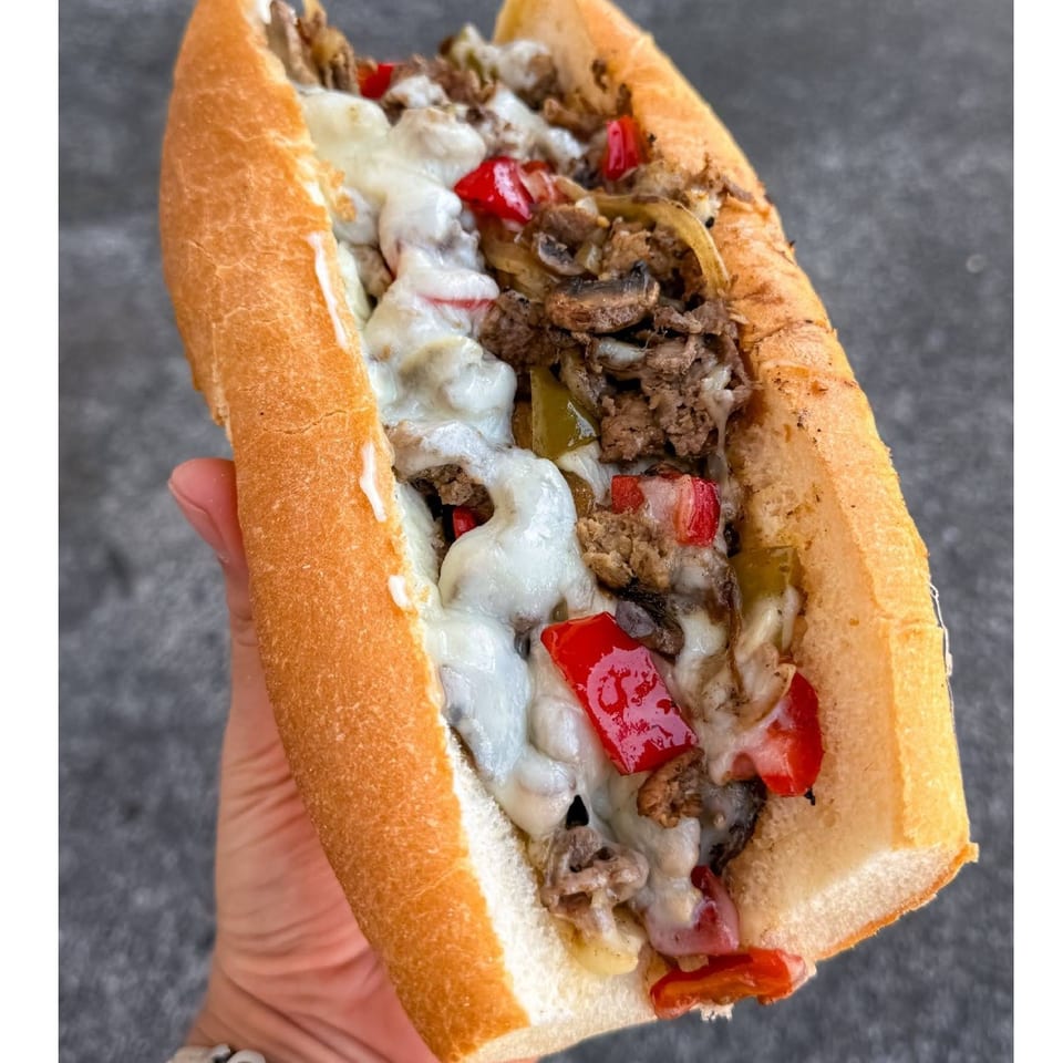 Hand holding a cheesesteak sandwich filled with sliced beef, melted cheese, mushrooms, onions, and red and green peppers.